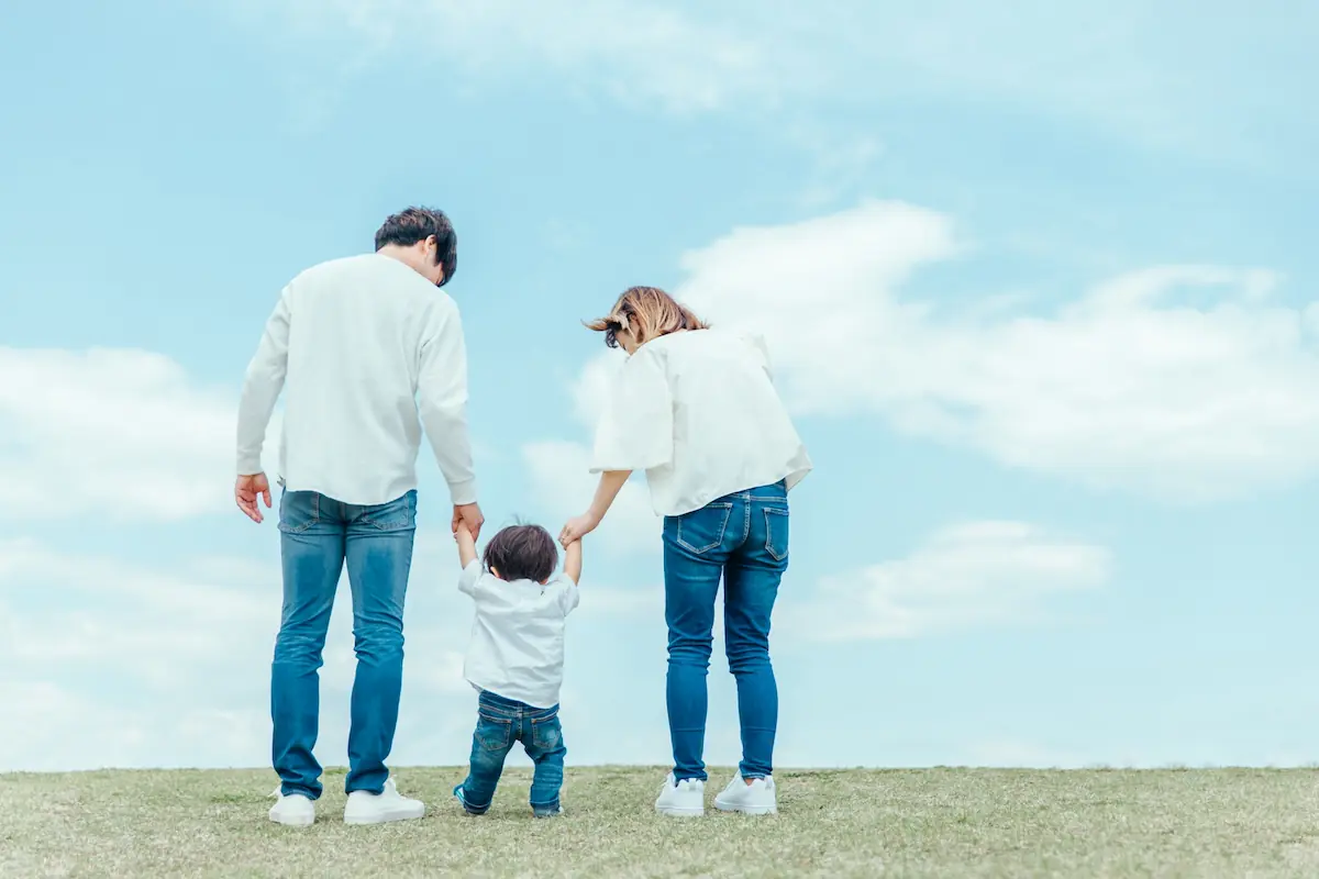 青空の下、家族3人が手をつないで歩く後ろ姿（子育て支援住宅のイメージ）